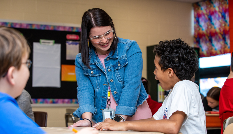 A teacher talking to students sitting at a desk while in a classroom.