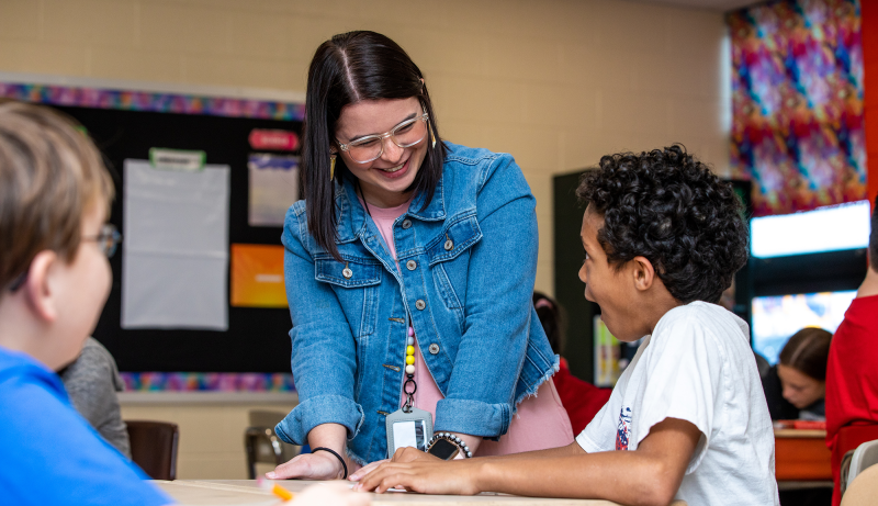  A teacher talking to students sitting at a desk while in a classroom. 